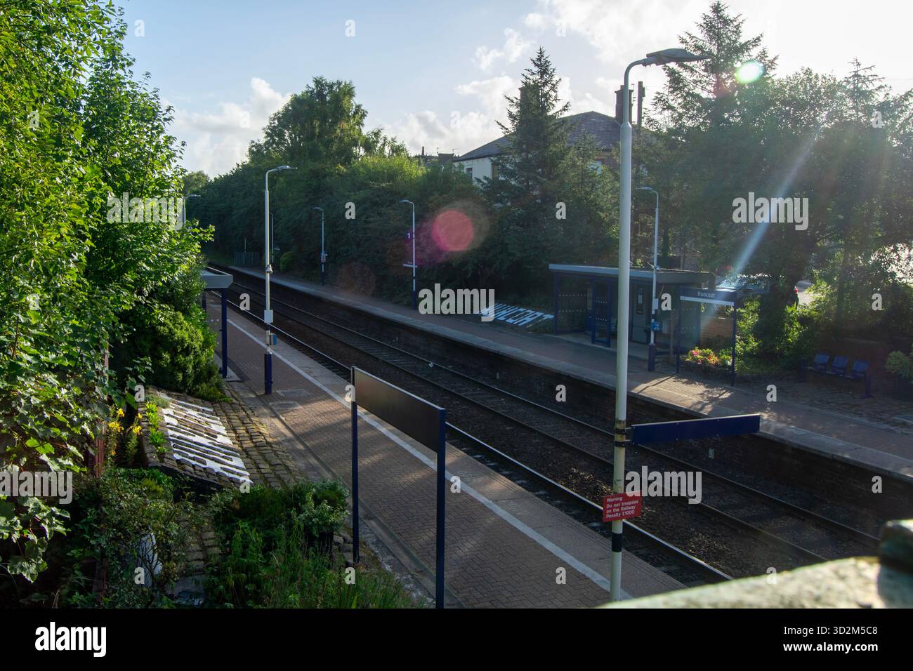 Una vista sopraelevata delle piattaforme e dei binari della stazione ferroviaria di Huncoat, bagnata dalla luce del sole, circondata da alberi lussureggianti ed edifici lontani. Foto Stock