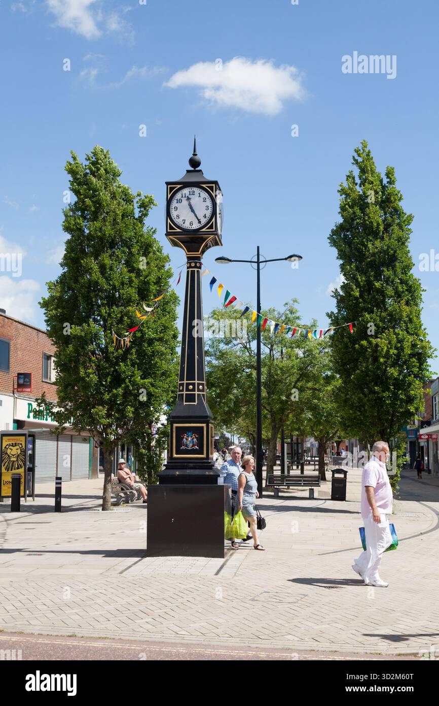 Una classica torre dell'orologio sorge in una vivace piazza della città sotto un cielo azzurro, con i bunting festivi e i pedoni che si godono una luminosa giornata estiva vicino a sh Foto Stock