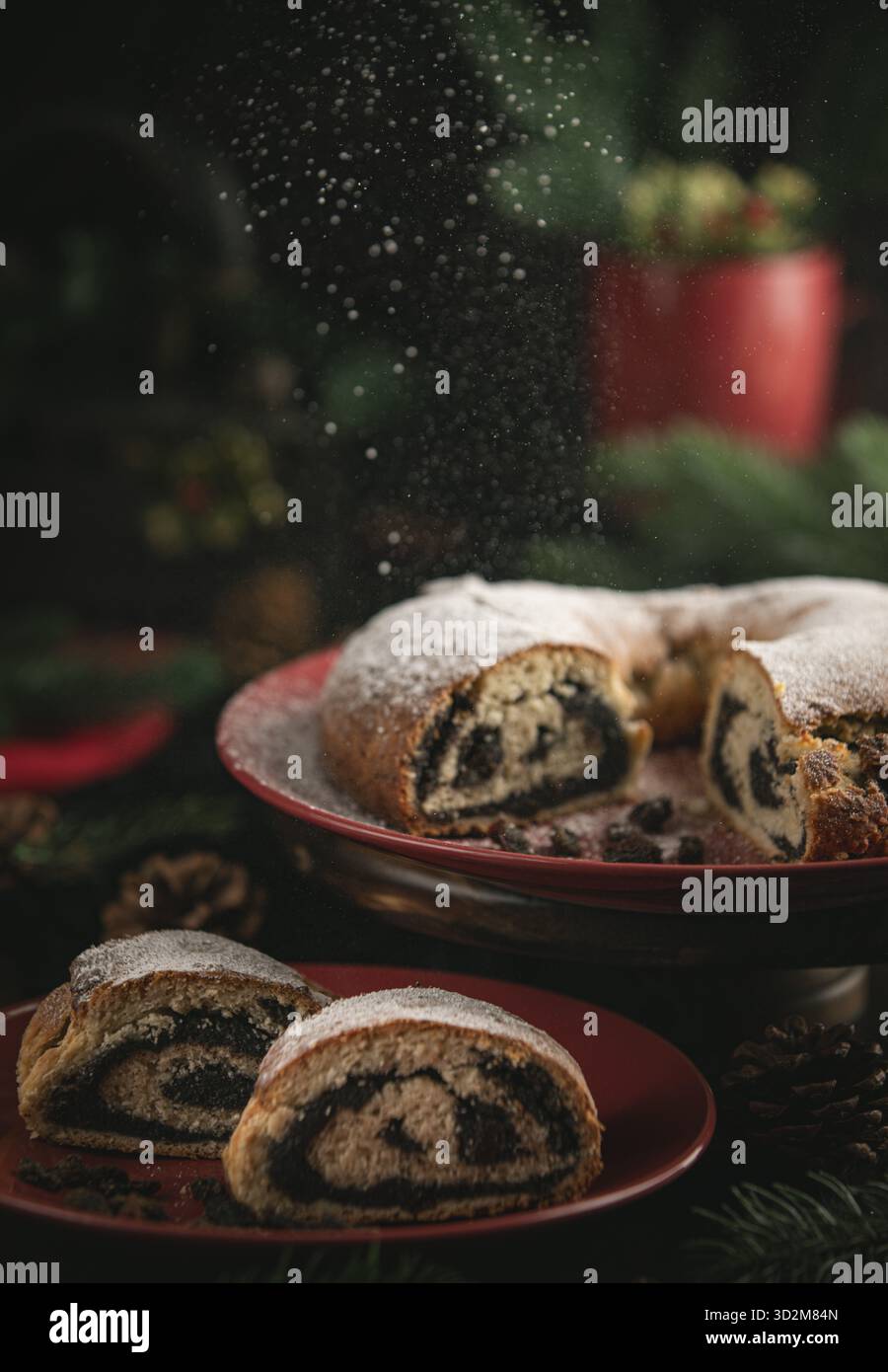 Lo zucchero a velo cade sulla torta di semi di papavero su un piatto rosso circondato da decorazioni natalizie Foto Stock