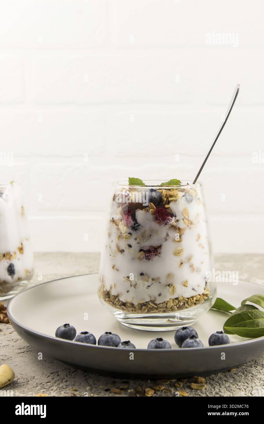 Un bicchiere di yogurt leggero, muesli, frutti di bosco freschi, noci si staglia su un piatto di ceramica con mirtilli e foglie verdi. colazione dietetica Foto Stock