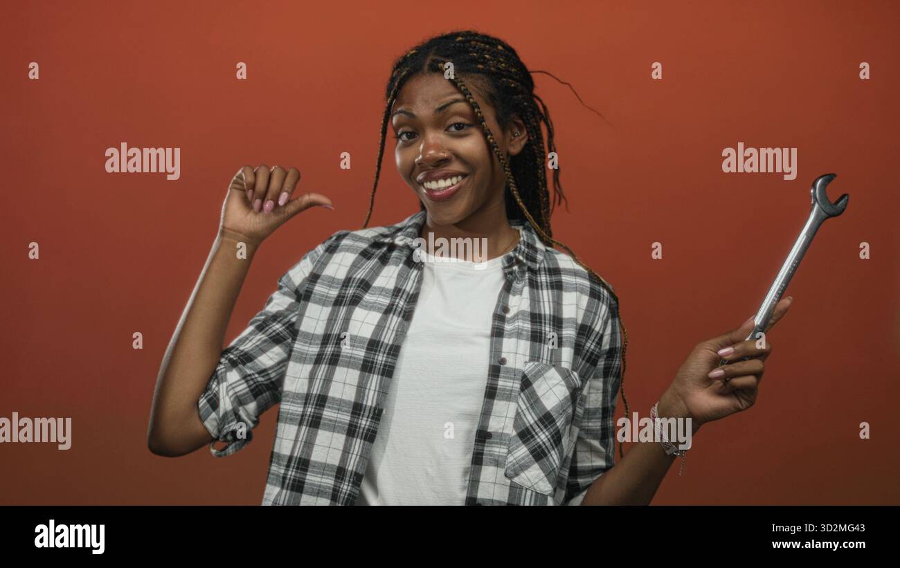 Donna che tiene la chiave e punta il pollice verso se stessa in studio, indossa una camicia a quadri e una T-shirt bianca con capelli intrecciati sullo sfondo arancione; autosufficiente p Foto Stock