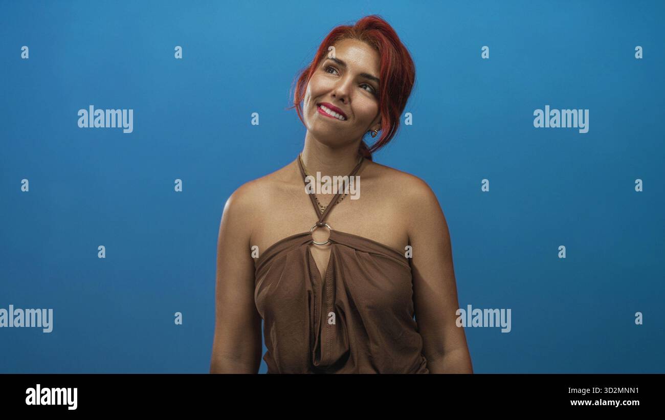 Donna con spalle esposte che sorride e testa inclinabile in studio con parete blu, capelli rossi e parte superiore marrone; giocosa disinvoltura. Foto Stock