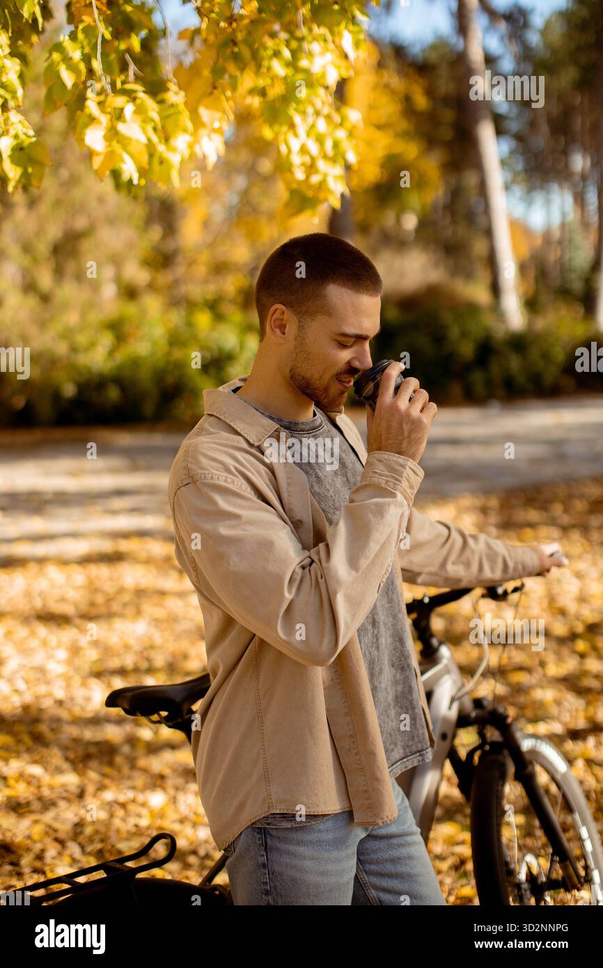 Un giovane beve un caffè mentre si trova accanto alla sua bicicletta in un bellissimo ambiente autunnale, circondato da foglie colorate. Foto Stock