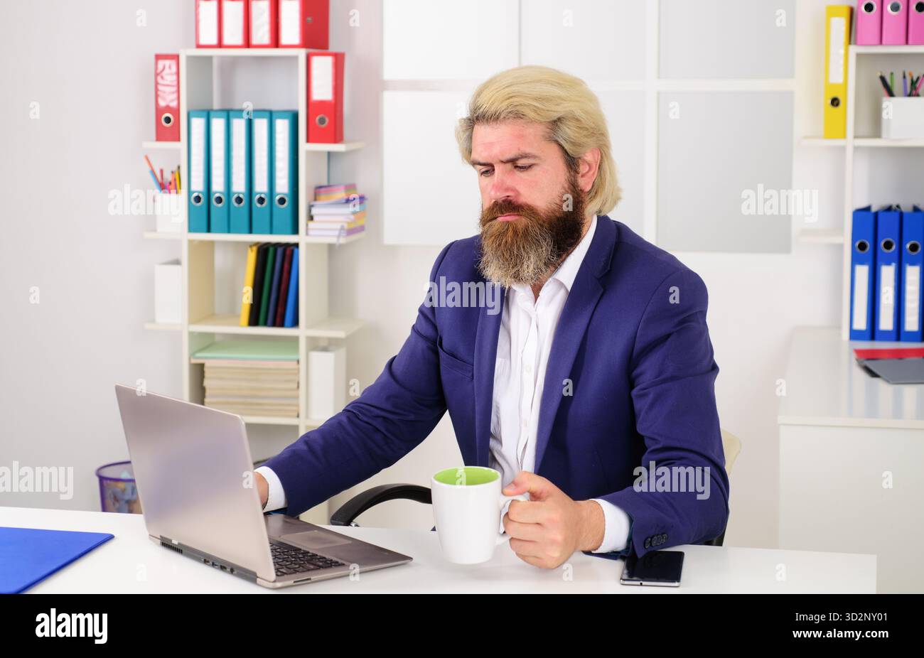 Uomo barbuto in tuta con una tazza di caffè che lavora su un progetto online su un computer portatile alla scrivania in ufficio. Serio uomo d'affari che beve caffè sul posto di lavoro. Sicuro di sé Foto Stock