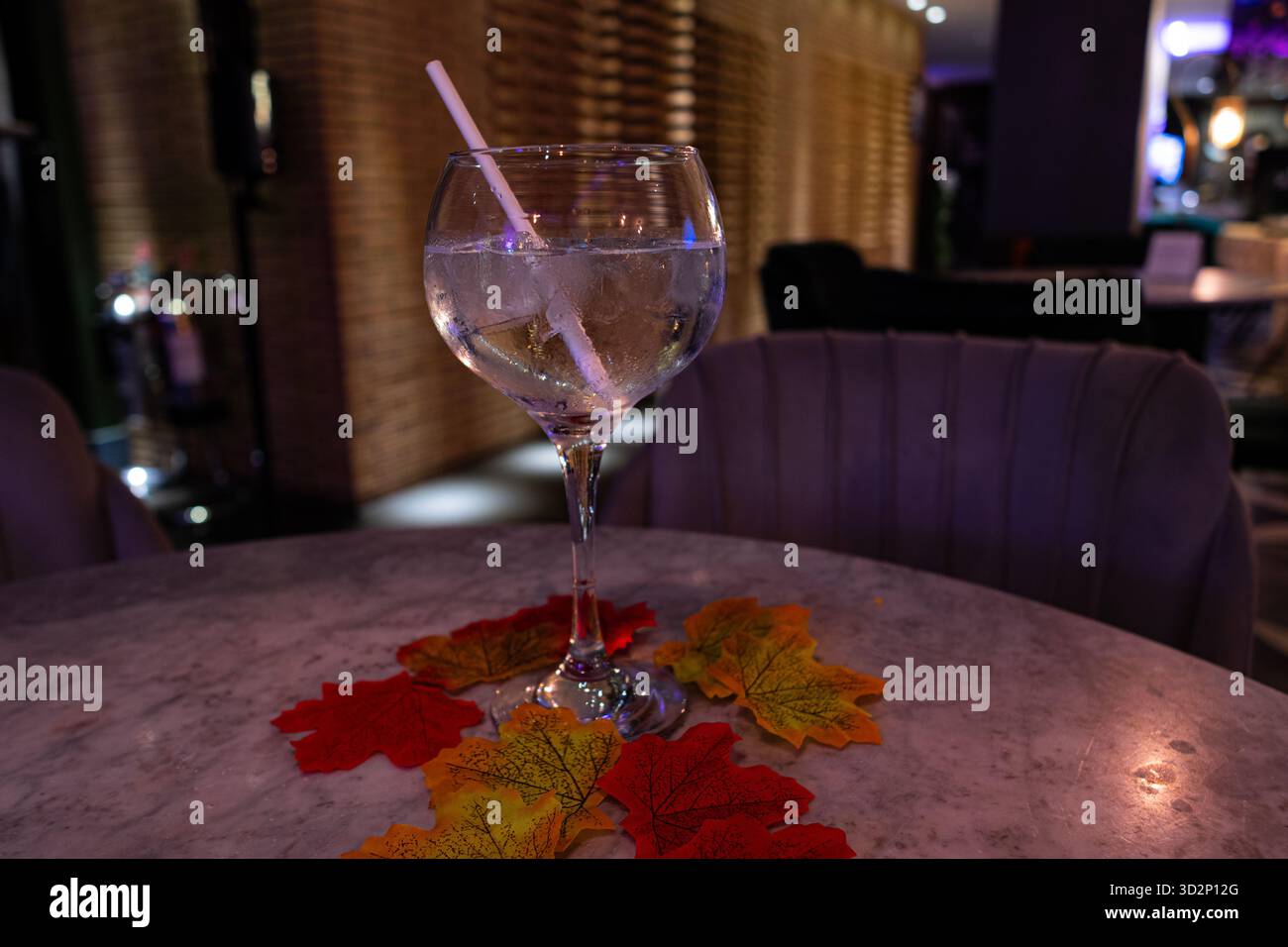 Una bevanda rinfrescante con ghiaccio e paglia, circondata da foglie autunnali colorate su un tavolo di marmo in un ambiente caldo e illuminato. Foto Stock