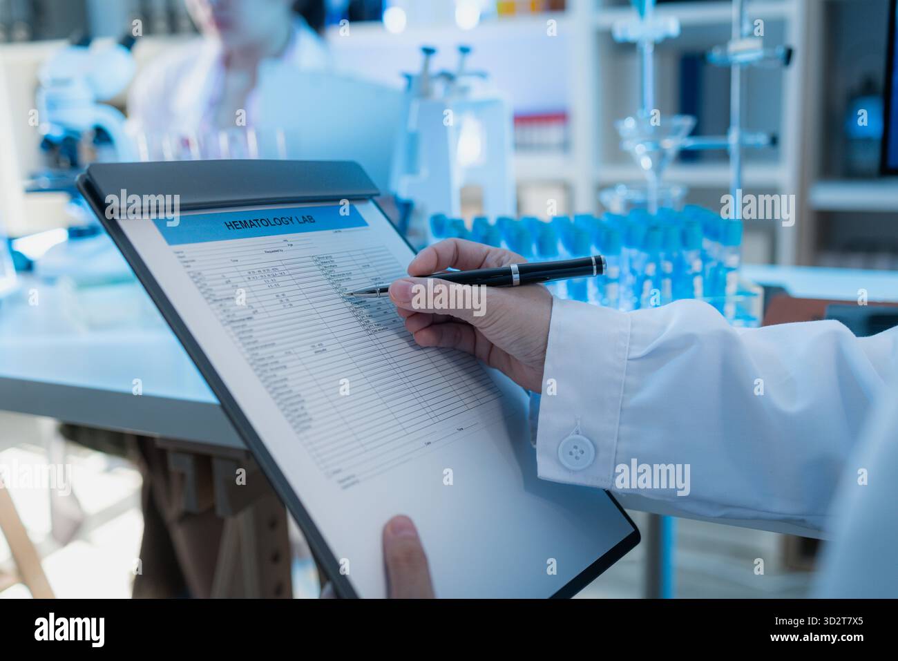 Vista ravvicinata di uno scienziato che esamina i dati di un laboratorio ematologico su una clipboard in un ambiente di laboratorio moderno. La scrivania è piena di bicchieri, tes Foto Stock