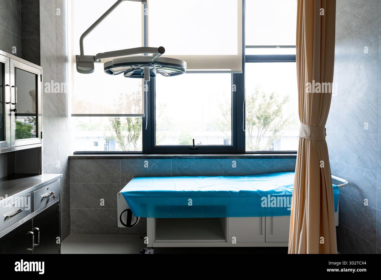 Una stanza per esami medici pulita con un letto rivestito di blu, una lampada chirurgica e una grande finestra con una tenda. Foto Stock