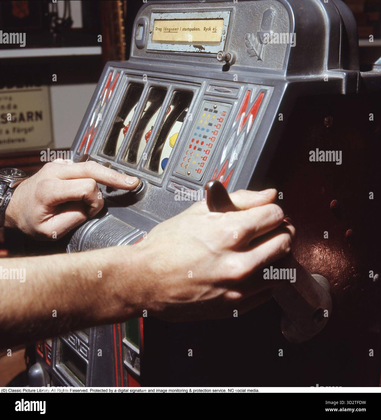 Sulla slot machine 1972. Una persona raffigurata nella slot machine. Una macchina da gioco in cui metti una moneta e tiri una leva che inizia a girare tre ruote. Una volta che si fermano e il set di ruote mostra lo stesso simbolo, vinci. Il bandito con un solo braccio, una classica slot machine, emerse alla fine del XIX secolo, con Liberty Bell di Charles Fey come pioniere nel 1895. Chiamato per la sua leva singola, che i giocatori tiravano per far girare i rulli, è diventato un punto fermo del casinò. Negli anni '1950, queste macchine meccaniche presentavano simboli colorati come frutta e campane, offrendo premi in denaro per combinazioni abbinate. Il Foto Stock