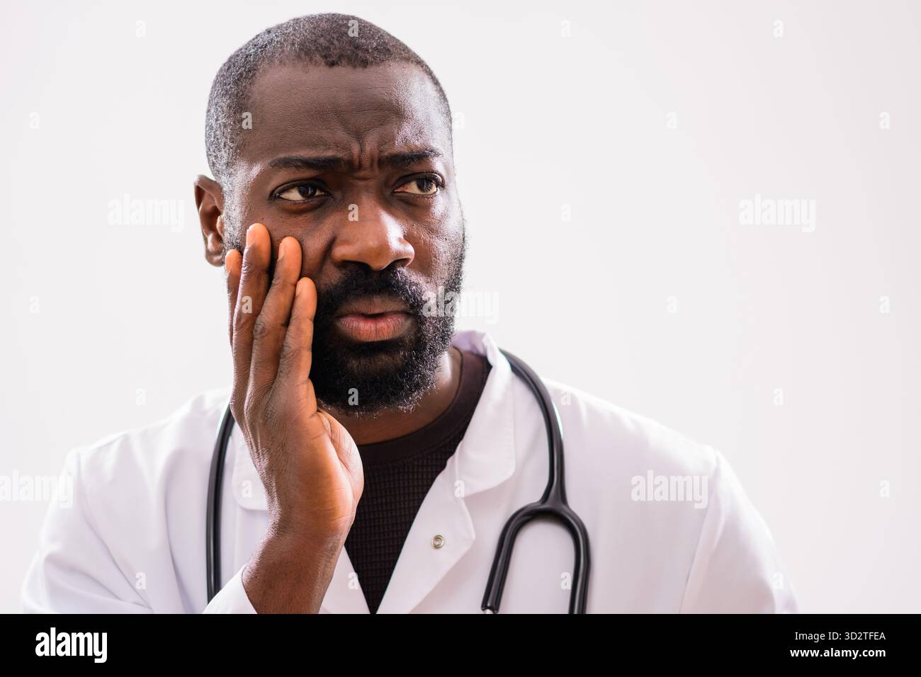 Ritratto di un medico afroamericano affaticato che sembra pensivo, mostrando segni di stress emotivo e stanchezza in un ambiente grigio da studio. Foto Stock