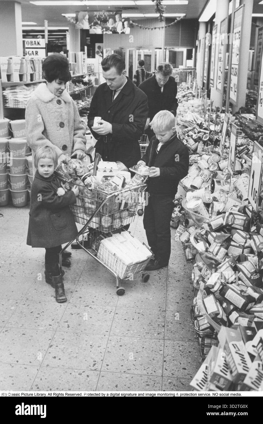 Shopping 1964. Interno di un negozio Co-op dove una famiglia con bambini sta facendo un grande negozio. Gli scaffali sono carichi di scatole e altri pacchetti, e la segnaletica evidenzia i prezzi allettanti del negozio. La moglie indossa un cappotto adatto al periodo e un elegante cappotto, mentre l'uomo sfoggia un trenchcoat elegante. Sia l'idea di self service che il carrello della spesa provenivano dall'America e miravano a ridurre le vendite. Il cliente poteva scegliere la spesa, mettere il carrello della spesa e procedere alla cassa del negozio. Foto Stock