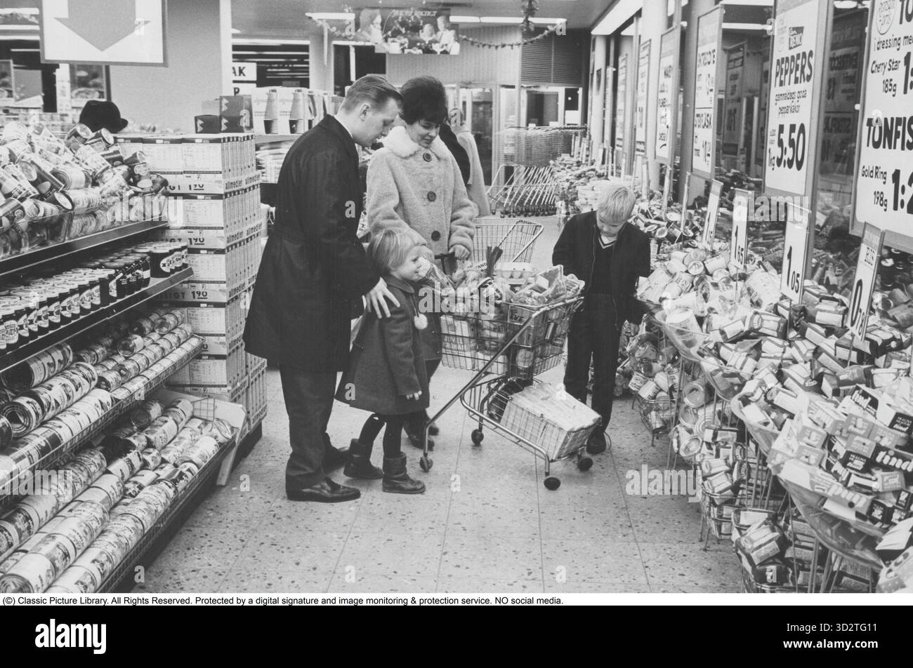 Shopping 1964. Interno di un negozio Co-op dove una famiglia con bambini sta facendo un grande negozio. Gli scaffali sono carichi di scatole e altri pacchetti, e la segnaletica evidenzia i prezzi allettanti del negozio. La moglie indossa un cappotto adatto al periodo e un elegante cappotto, mentre l'uomo sfoggia un trenchcoat elegante. Sia l'idea di self service che il carrello della spesa provenivano dall'America e miravano a ridurre le vendite. Il cliente poteva scegliere la spesa, mettere il carrello della spesa e procedere alla cassa del negozio. Foto Stock