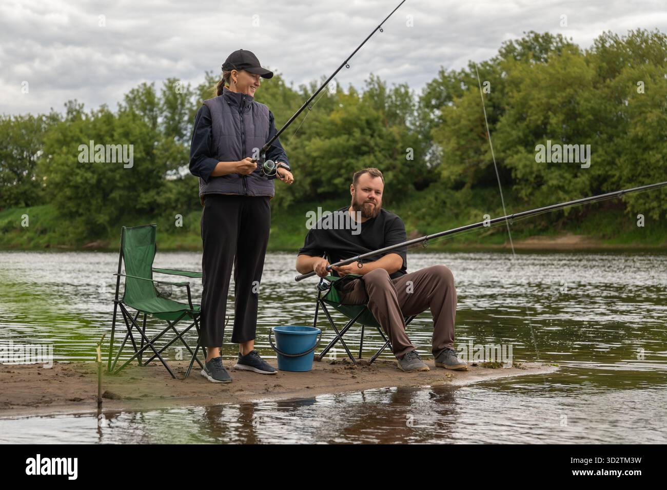 Uomo e donna che pescano sulla riva del fiume, immergendosi nella quiete circostante mentre vi godete una rilassante attività all'aperto insieme. Sedie da campeggio e un Foto Stock