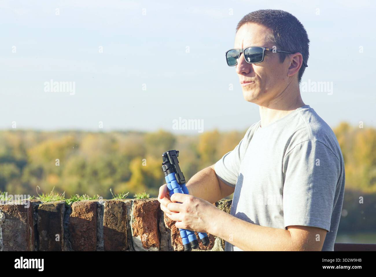 Uomo che tiene in mano un piccolo treppiede flessibile mentre si gode una vista panoramica autunnale da un punto panoramico. La prospettiva turistica cattura e vlogging stile di vita Foto Stock