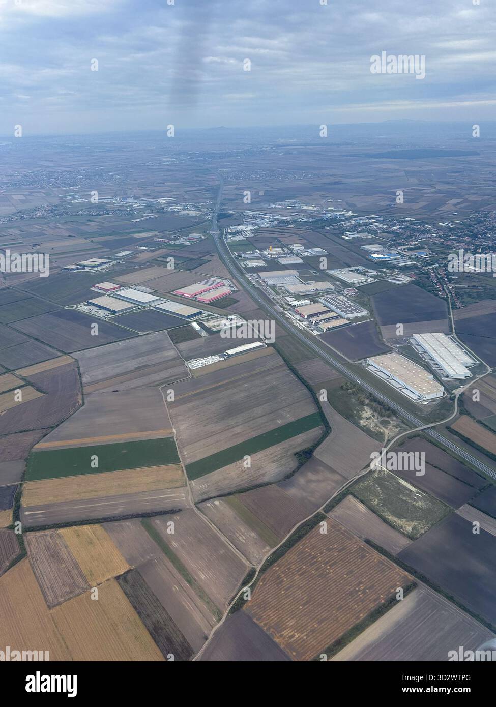 Foto aerea di vasti terreni agricoli con campi geometrici e moderni magazzini industriali sotto un cielo nuvoloso, che mostrano il contrasto rurale e urbano. - Immagine stock catturata con smartphone