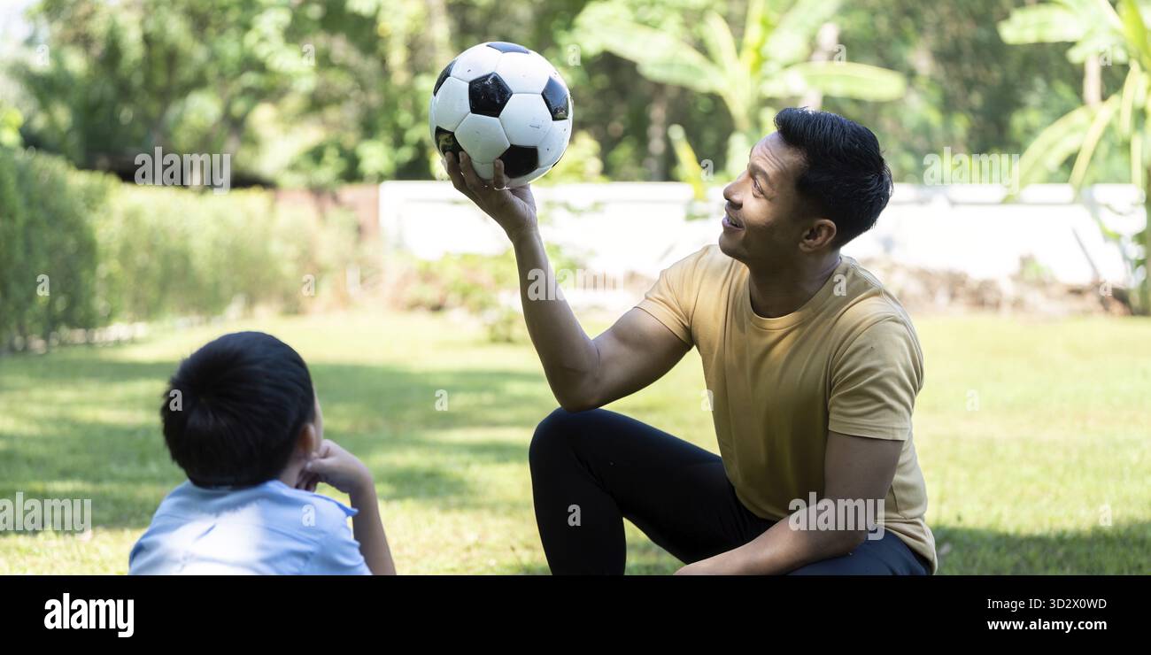 Un padre che insegna giocosamente a suo figlio abilità calcistiche, incoraggiando il benessere e il lavoro di squadra nel loro giardino all'aperto, pieno di gioia e risate Foto Stock