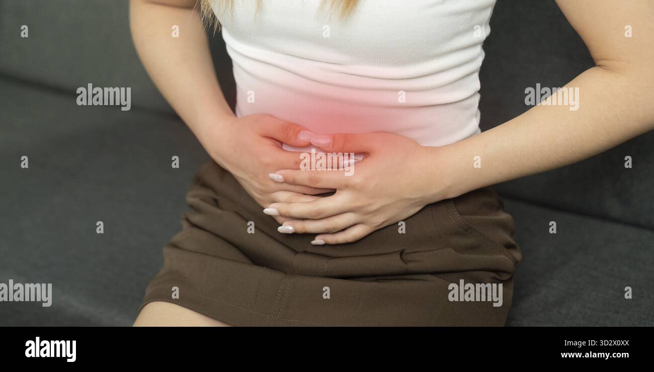 Una donna seduta su un divano, che afferra il suo addome con disagio. Rappresenta dolore addominale e consapevolezza della salute Foto Stock