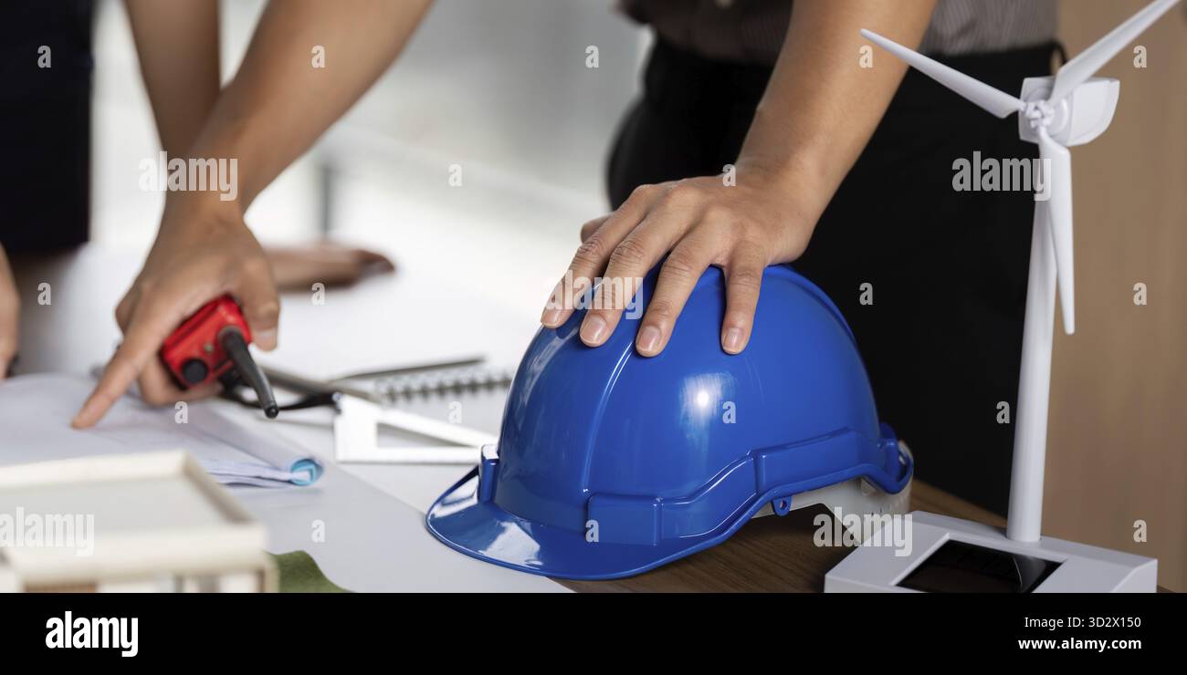 Un ingegnere che utilizza strumenti e un elmetto blu mentre pianifica soluzioni innovative per l'energia sostenibile con modelli e progetti su un tavolo Foto Stock