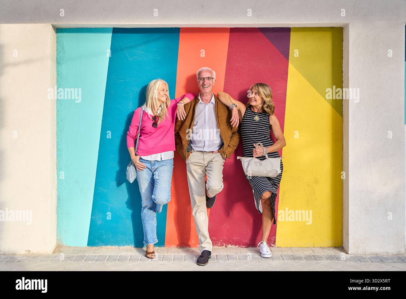 Tre amici anziani sorridenti appoggiati su una vivace parete dipinta, godendosi il loro felice stile di vita in pensione all'aperto in una giornata di sole Foto Stock