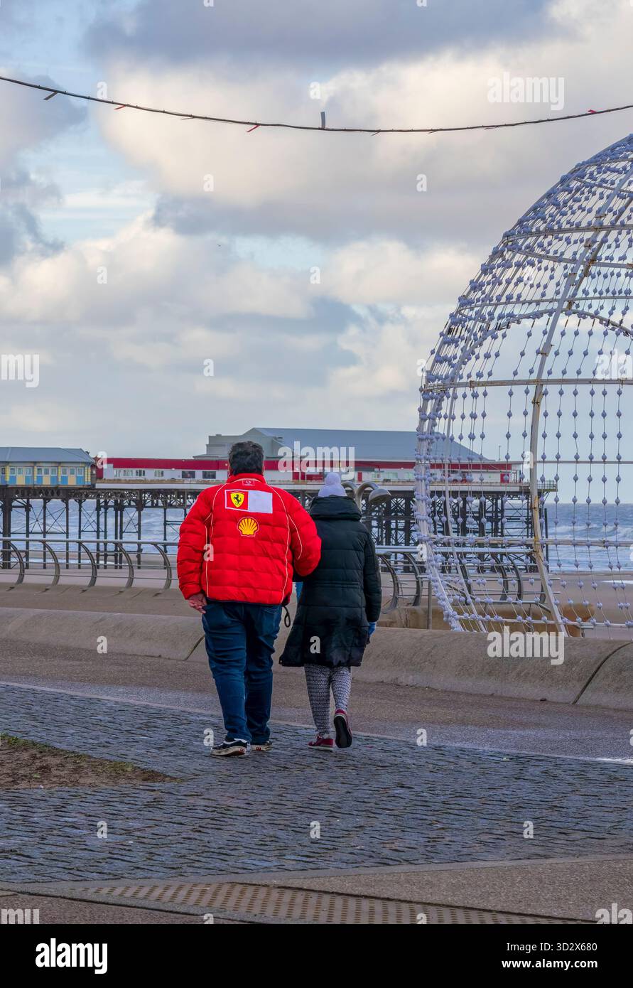 Un uomo e una donna camminano, braccio in braccio, sul lungomare di Blackpool, nel Regno Unito. Uno dei tre moli di Blackpool, Central Pier, può essere visto sullo sfondo Foto Stock
