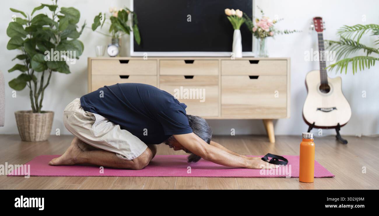 Un uomo anziano che esplora le tecniche di benessere praticando posizioni di yoga a casa, concentrandosi sulla salute e il relax Foto Stock