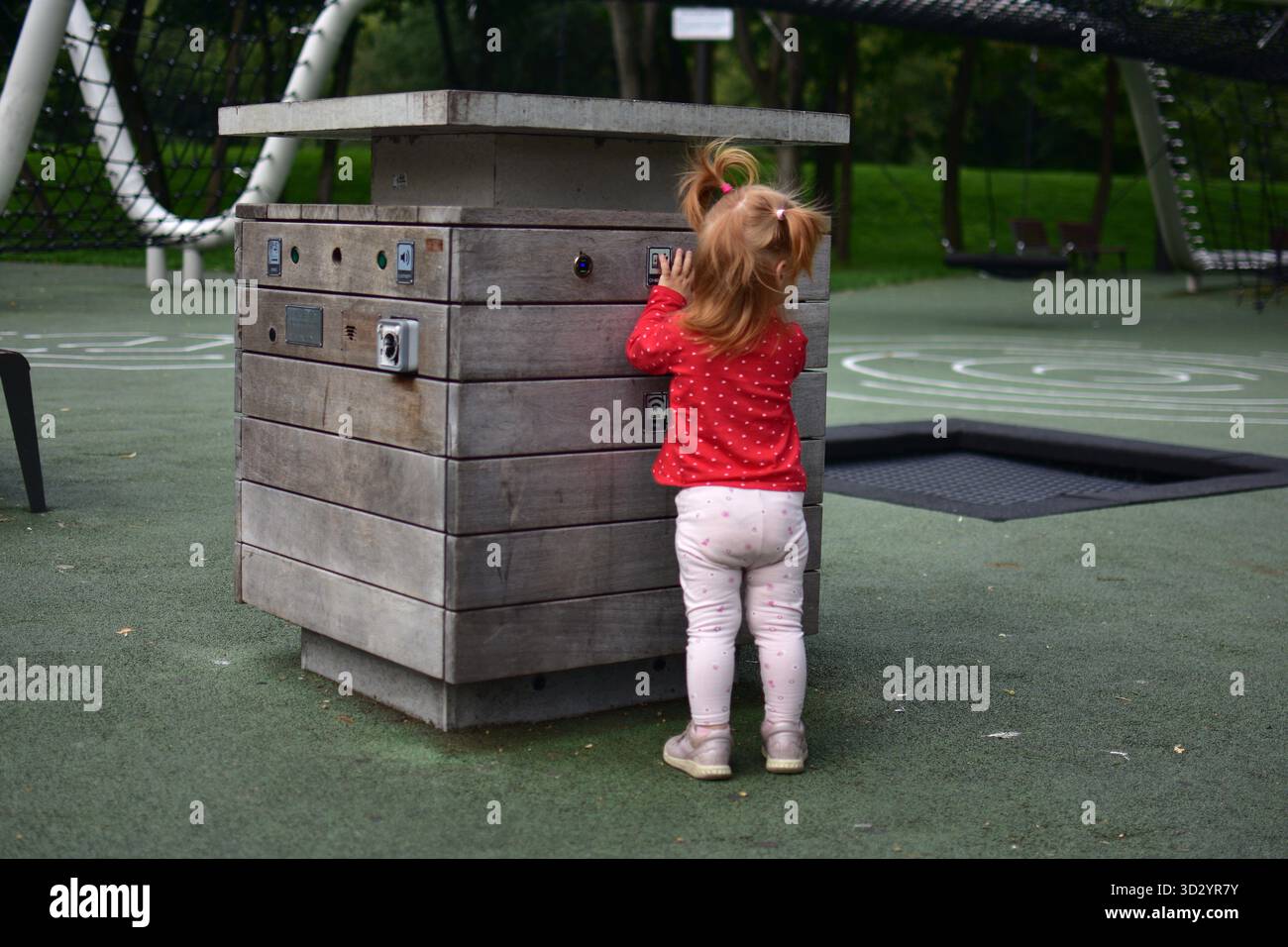 Bambina in piedi al box giochi interattivo in legno premendo i pulsanti, curioso momento esplorativo, gioco all'aperto e apprendimento con il tocco Foto Stock