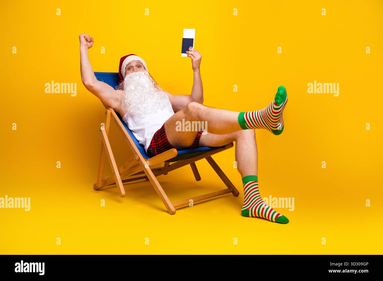 Babbo Natale si rilassa in un luminoso studio giallo posato su una sedia di legno mentre tiene in mano una tessera con calzini a righe e pantaloncini a quadri per una scena domestica di festa Foto Stock