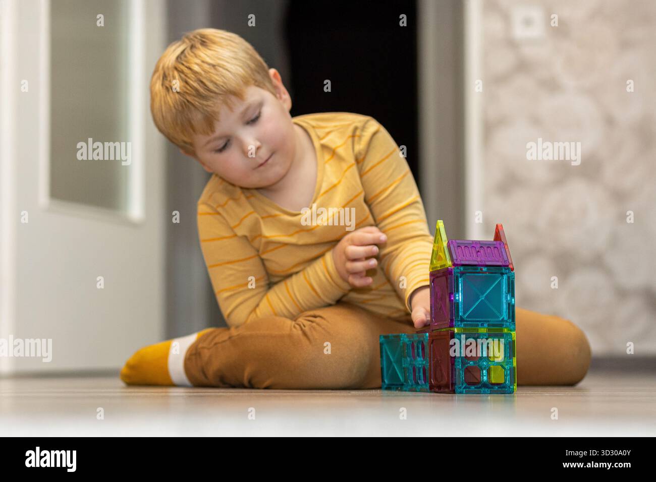 Un bambino si concentra sulla costruzione di una casa vibrante utilizzando piastrelle magnetiche. La camera è ben illuminata e presenta un decor semplice e casalingo. Foto Stock