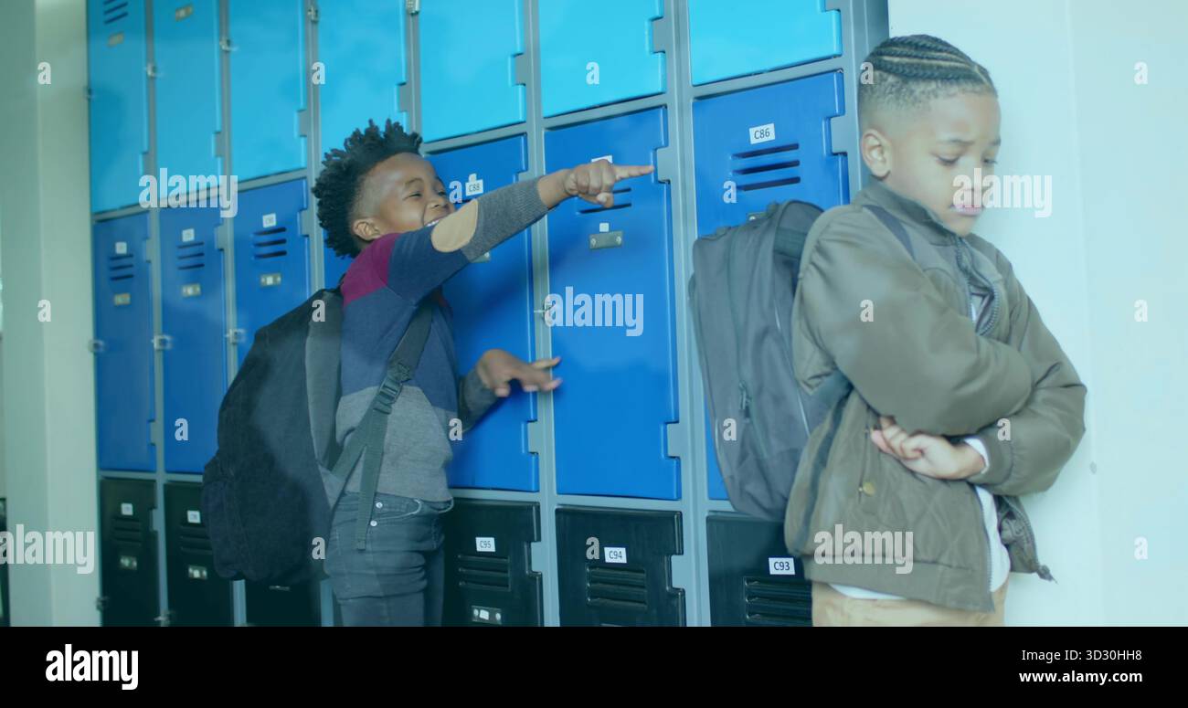 Ragazzo ridendo che punta, prende in giro il compagno di classe contro gli armadietti numerati nel corridoio scolastico, con zaini Foto Stock