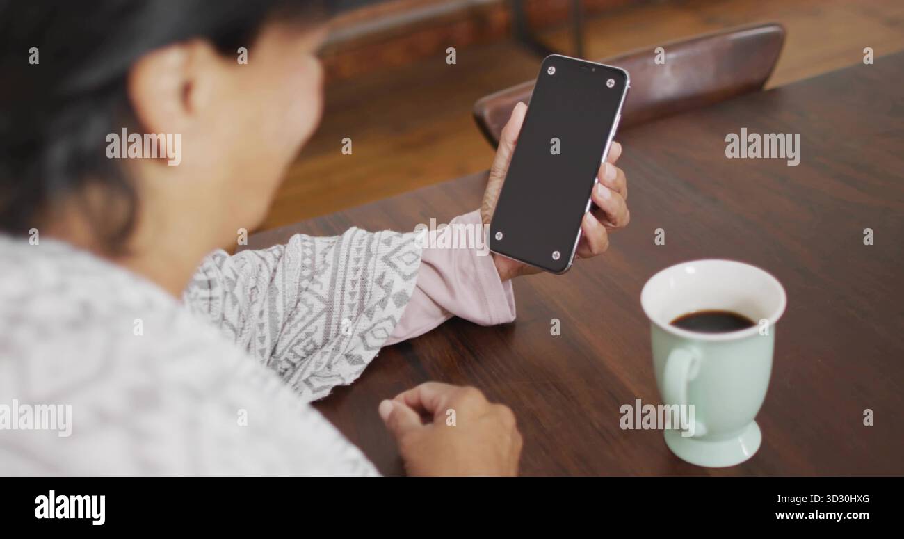 Tenendo in mano lo smartphone donna indiana anziana che esamina lo schermo vuoto al tavolo da pranzo di casa, con tazza da caffè Foto Stock