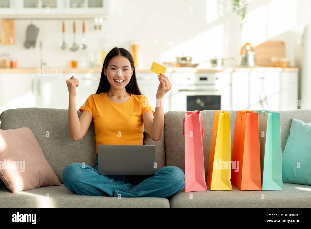 Eccitata donna asiatica che fa shopping online a casa con laptop e borse colorate Foto Stock