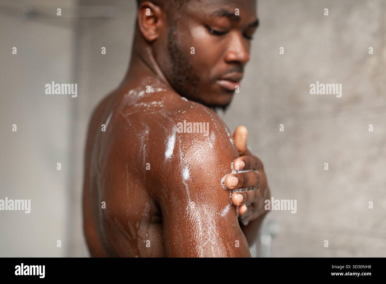 Uomo afroamericano muscolare che lava il corpo sotto la doccia con sapone e schiuma al chiuso Foto Stock
