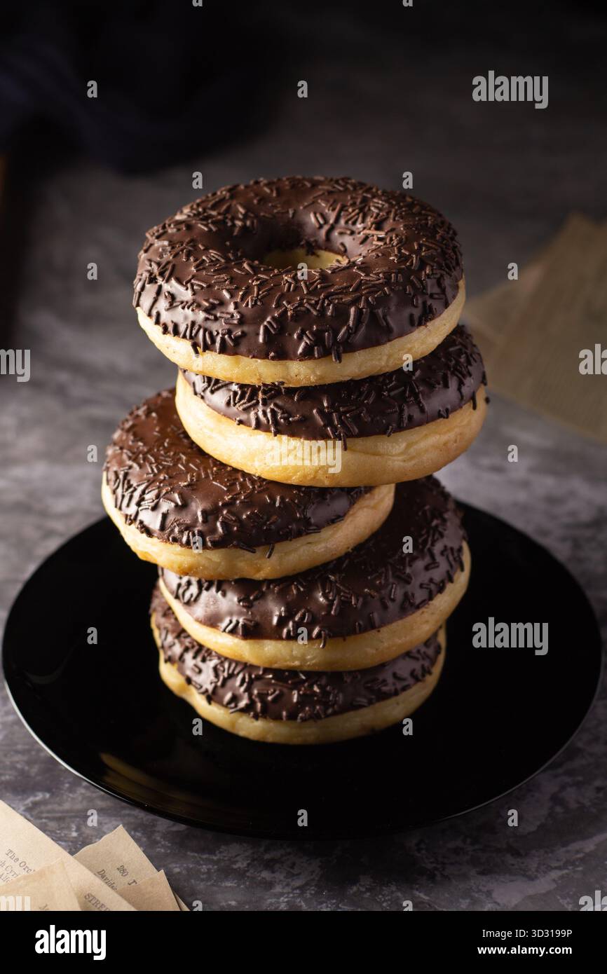 Una pila di cinque ciambelle smerigliate al cioccolato poggia su un piatto nero, mettendo in risalto il ricco cioccolato e gli sprinkles. Lo sfondo suggerisce un caldo, inviti Foto Stock