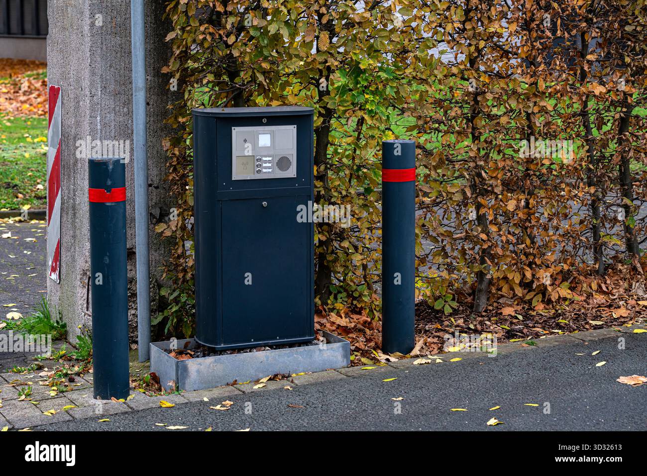 Un moderno citofono sul cancello sullo sfondo di vivaci foglie autunnali. Due pali con marcature rosse incorniciano l'interfono, evidenziando la c. Stagionale Foto Stock