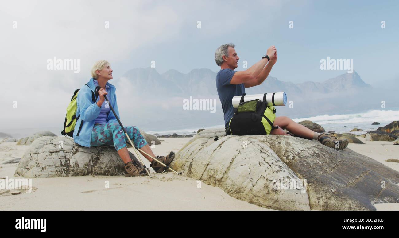 Coppia senior seduto che cattura la vista nebbiosa delle montagne sulle rocce costiere, con zaini Foto Stock
