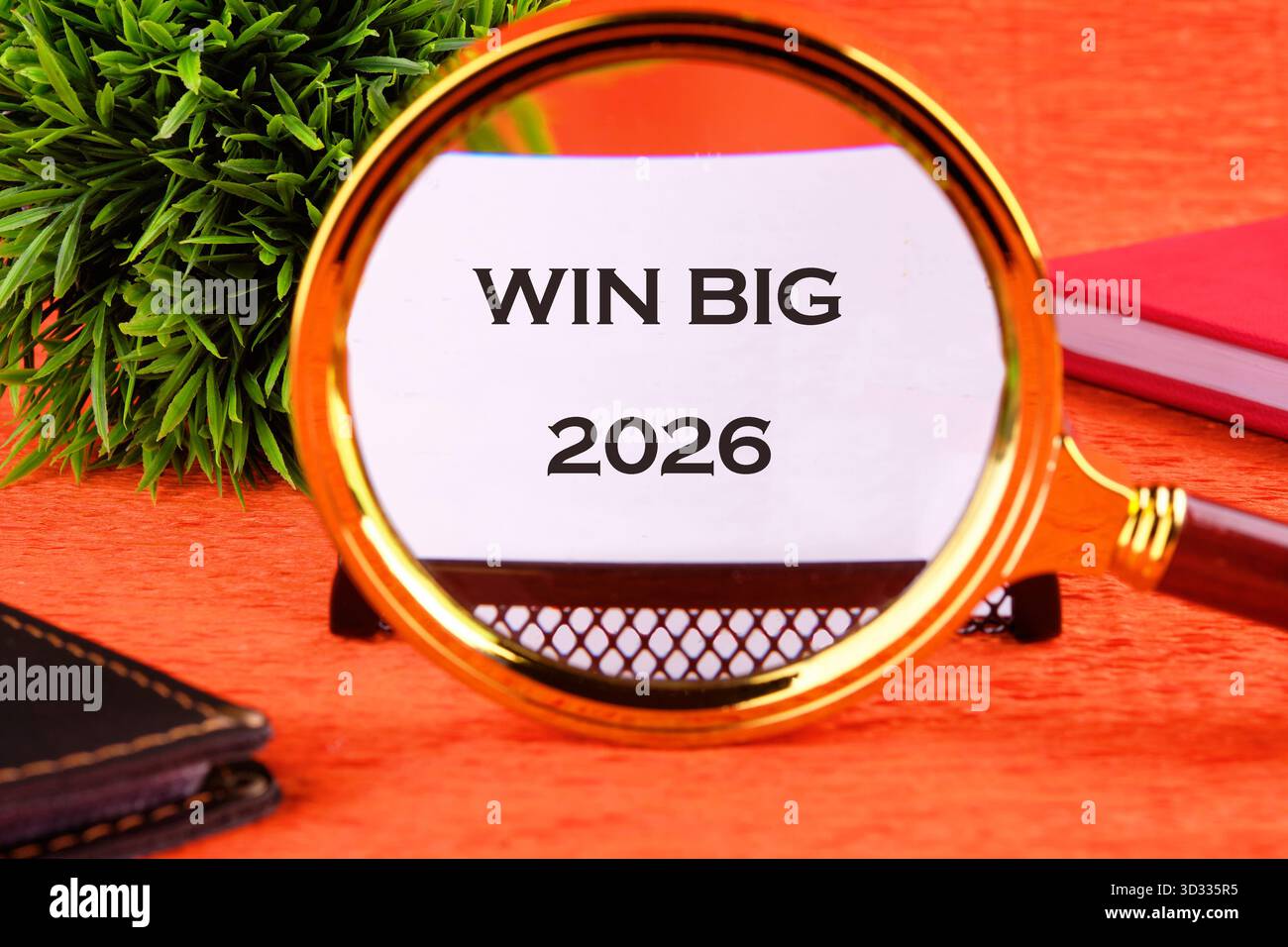Una lente di ingrandimento si concentra su un documento che dice Win Big 2026. Un quaderno nero e un libro rosso si trovano accanto al vetro su una superficie arancione. Foto Stock
