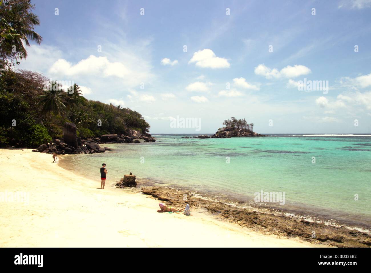 Una piccola insenatura di sabbia dorata si apre sul mare turchese di Mahé, Seychelles. Alcune persone si godono il sole e l'acqua limpida mentre una piccola isola verde Foto Stock