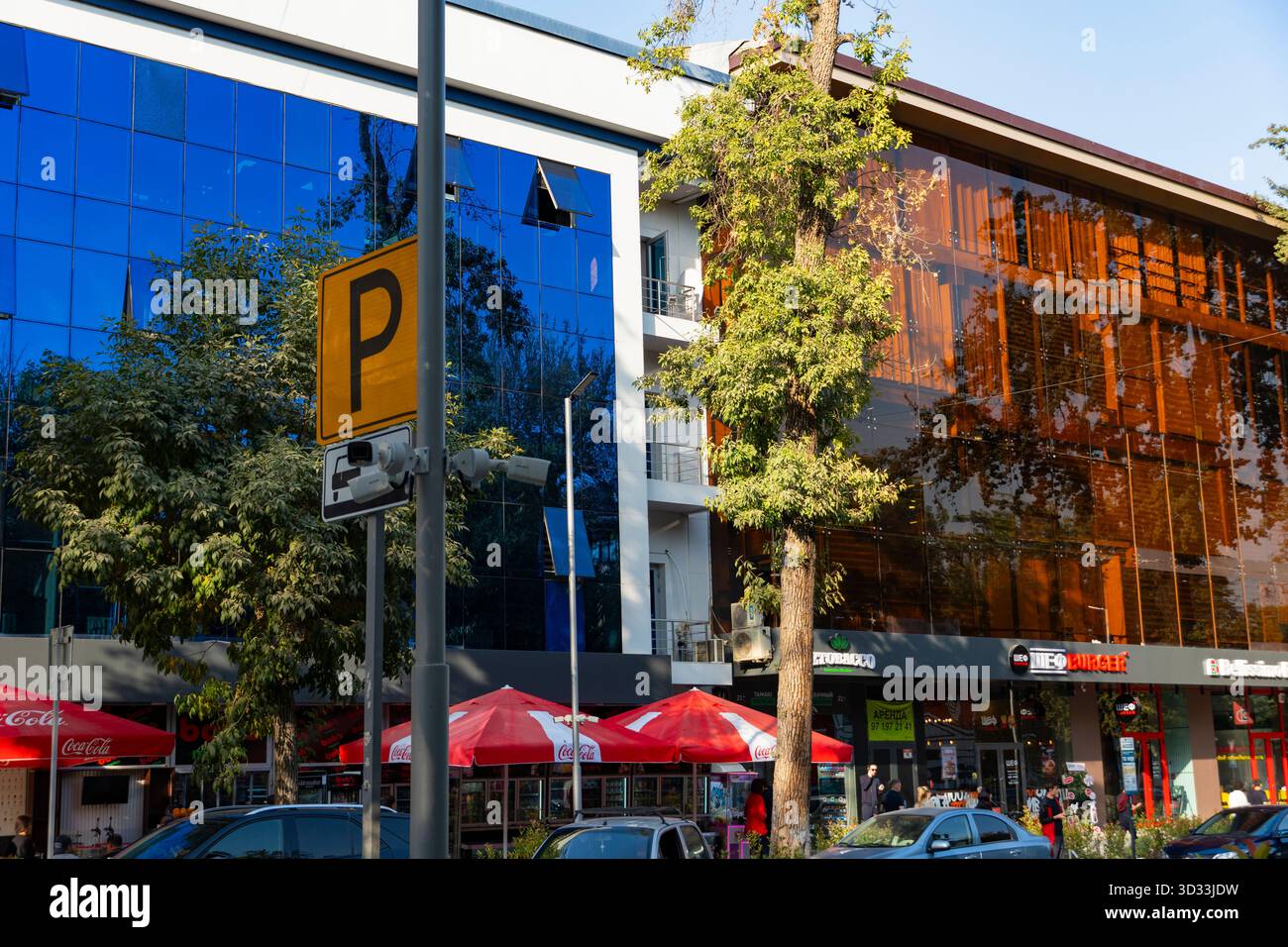 Architettura cittadina contemporanea con facciate in vetro, segnaletica di parcheggio, alberi e persone in una giornata di sole. Vista sulla strada urbana con riflessi e caffetterie Foto Stock
