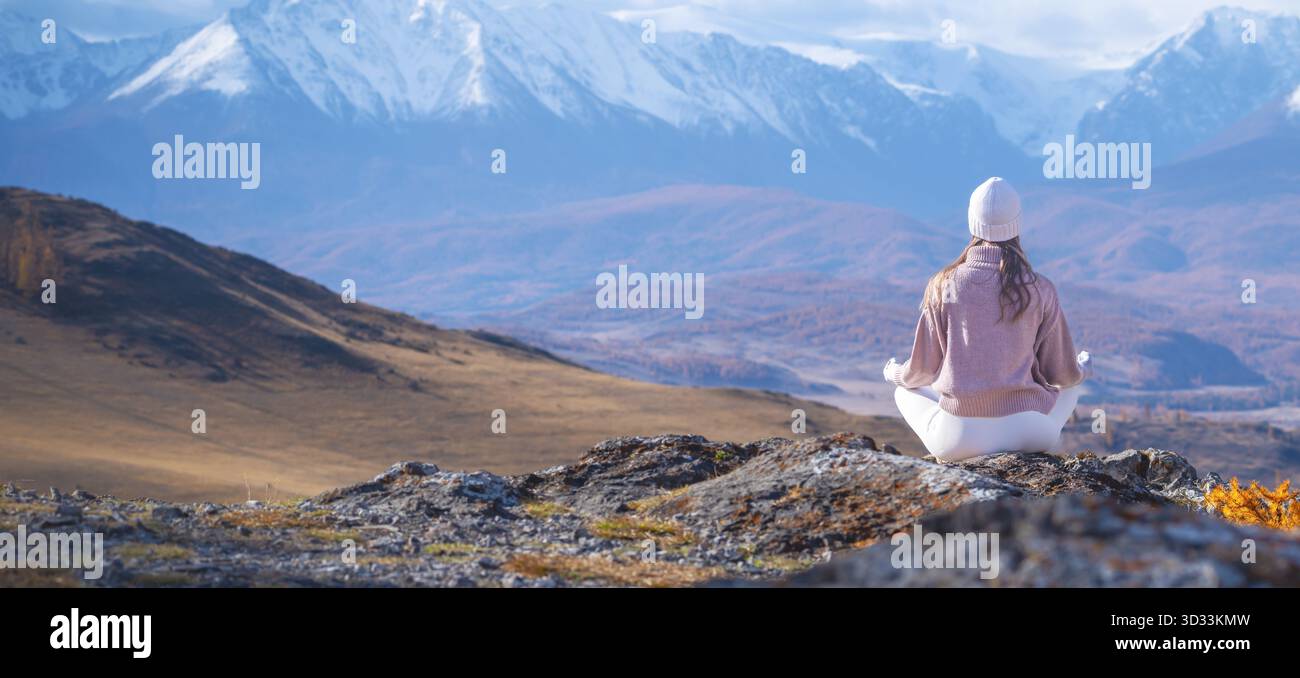Una donna siede pacificamente in meditazione sulla cima di un affioramento roccioso, circondato da montagne mozzafiato e paesaggi autunnali colorati. Abbraccia il tra della natura Foto Stock