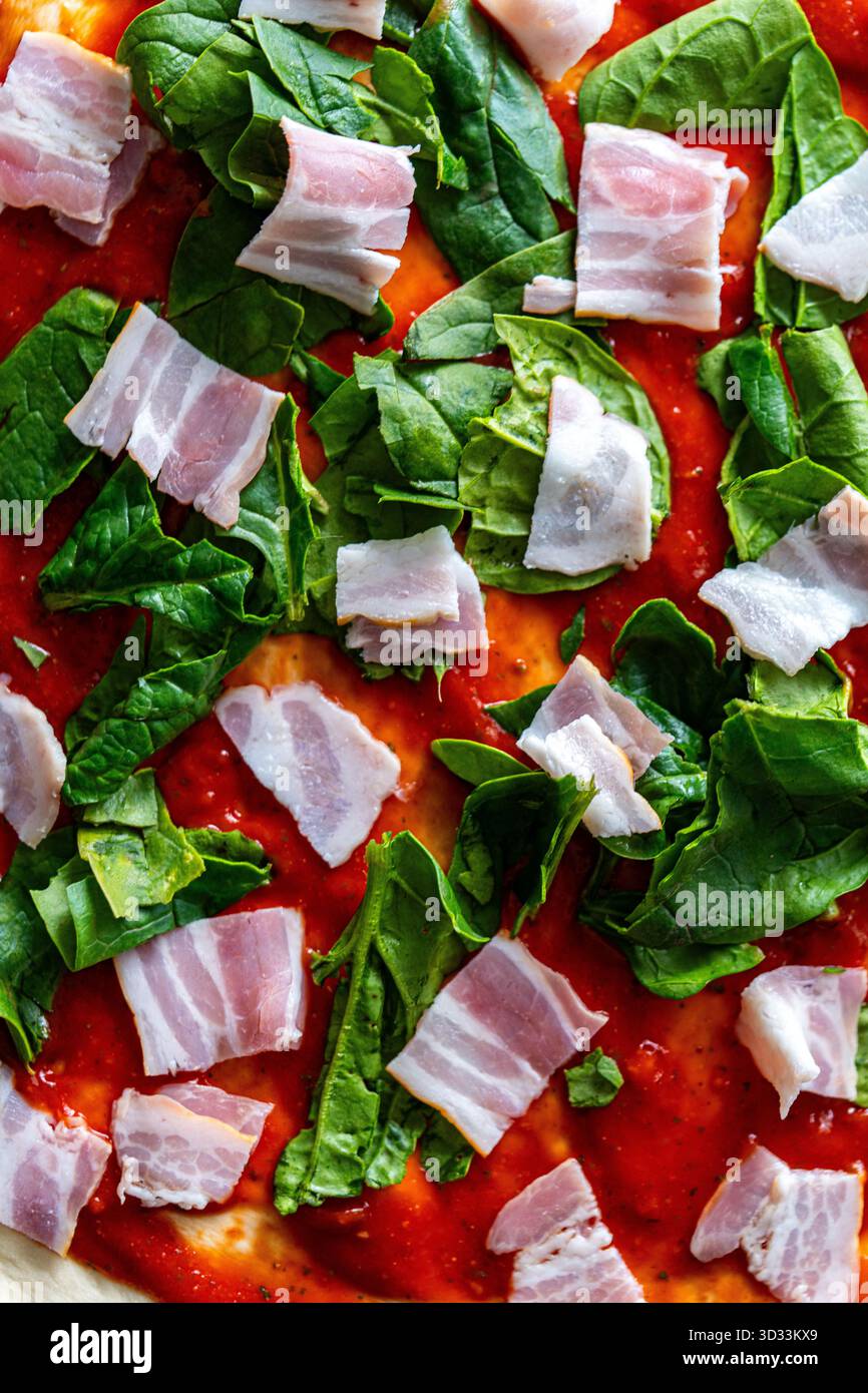 Pizza cruda con salsa di pomodoro, spinaci e pancetta. Primo piano, vista dall'alto, sfondo della consistenza della pizza al pomodoro cruda. Foto di alta qualità Foto Stock
