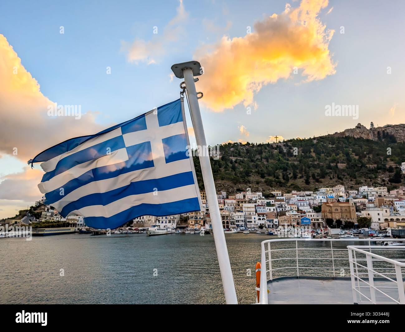 Bandiera greca che vola su Un traghetto con Pothia e il porto sullo sfondo al tramonto, simbolo di viaggi e cultura Foto Stock