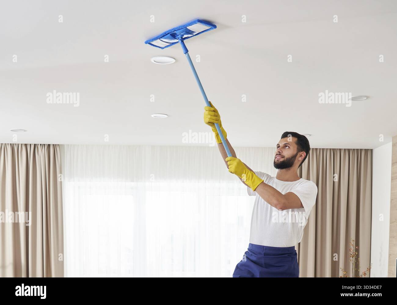 Uomo che tiene un mucchio di mocio, pulisce il soffitto in soggiorno Foto Stock
