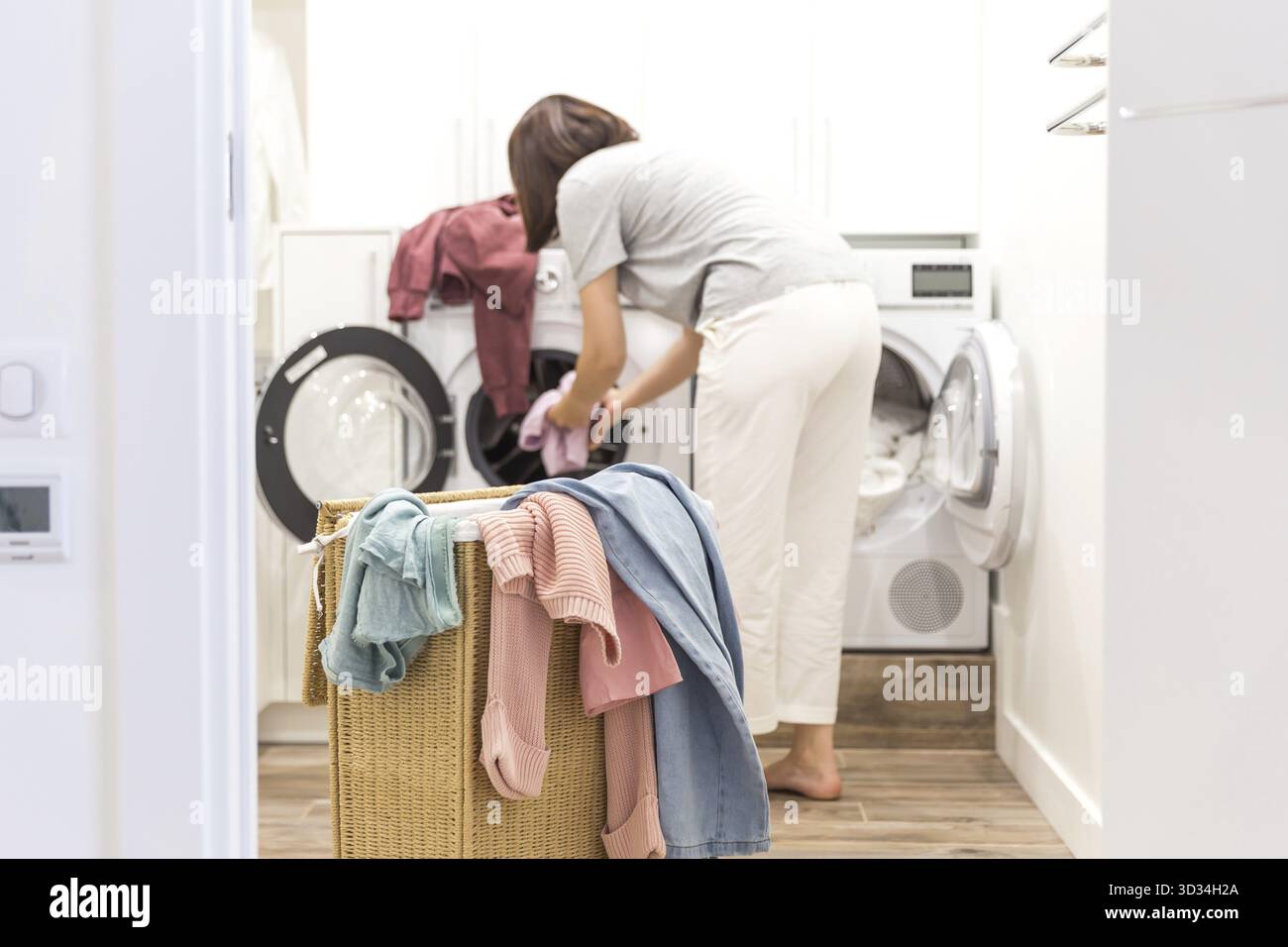 Una giovane donna che carica la lavatrice e il cestino pieno di vestiti sporchi nella lavanderia Foto Stock
