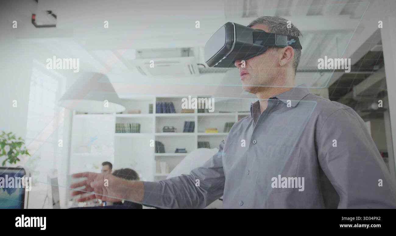 Uomo bianco maturo che indossa un visore per la realtà virtuale che raggiunge un ufficio aperto, con computer Foto Stock