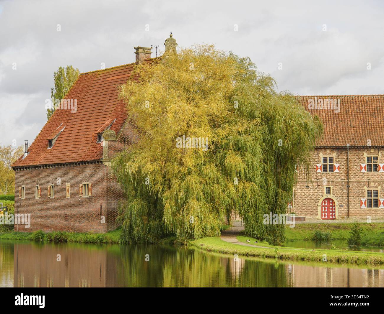 Casa in mattoni accanto allo stagno con tetto in piastrelle rosse e salice lussureggiante. Tranquilla scena rurale, Raesfeld, Muensterland, Germania Foto Stock