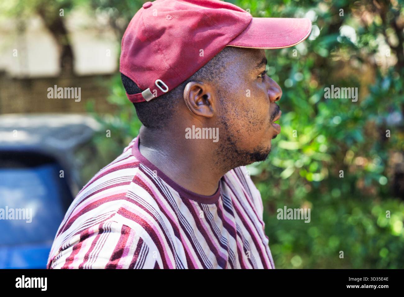 un giovane uomo africano in sovrappeso che cammina nel cortile, indossa una t-shirt a righe e un berretto Foto Stock