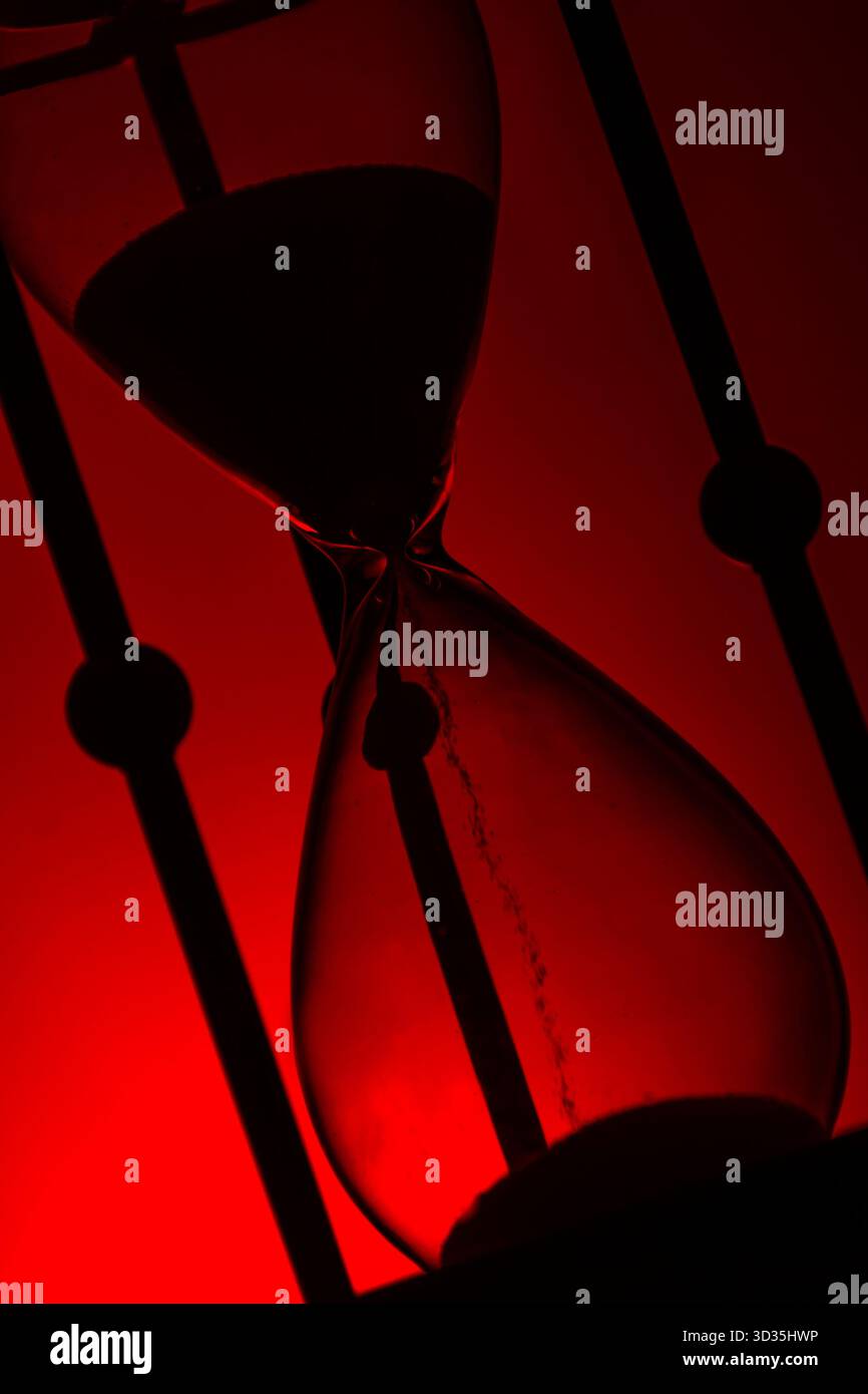 Primo piano di clessidra con sabbia che scorre, sagomata su un rosso brillante. Un simbolo drammatico di tempo, urgenza e riflessione filosofica. Foto Stock