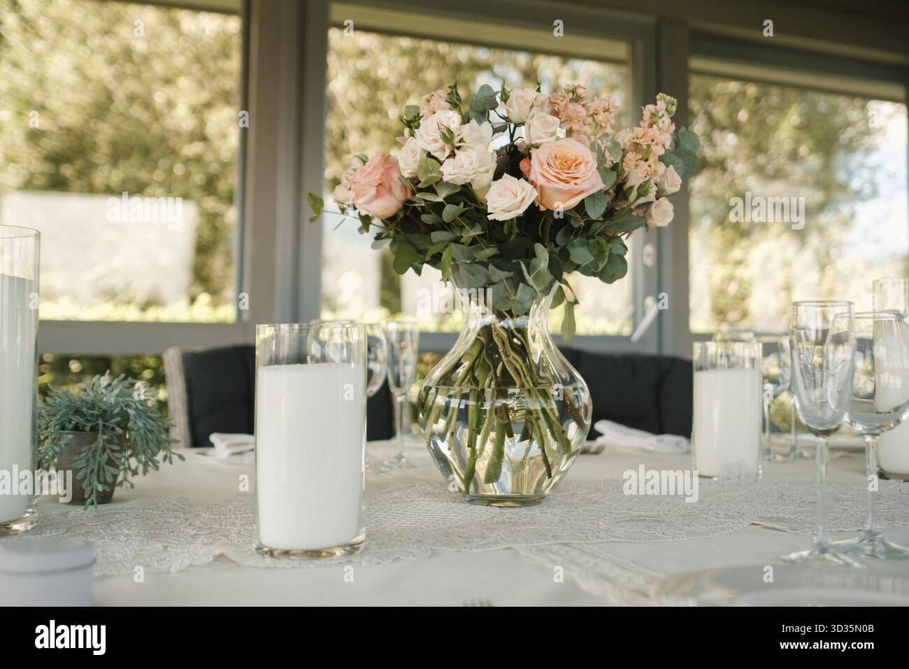 Un tavolo splendidamente organizzato presenta un vaso di vetro pieno di rose rosa e bianche, circondato da alti vetri e una tovaglia di pizzo, tutto illuminato Foto Stock