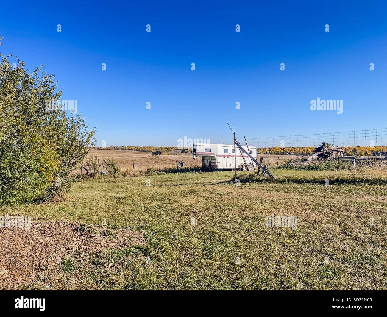 Ampia scena rurale caratterizzata da un vecchio rimorchio, recinzione intemprata e prateria aperta sotto un cielo blu brillante. Un ambiente tranquillo e isolato di campagna evoca soli - Immagine stock catturata con smartphone