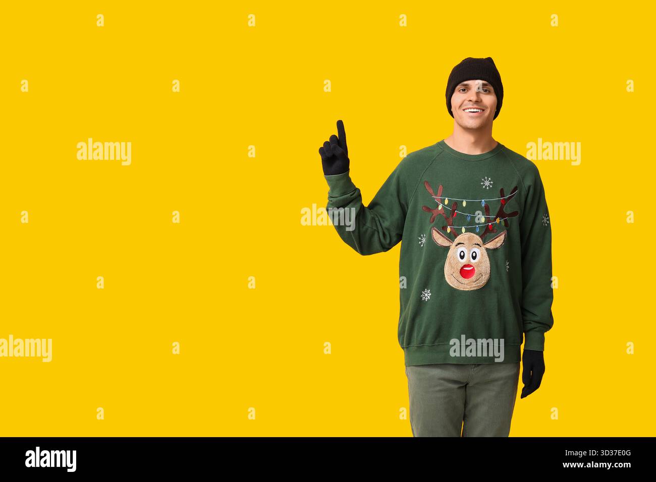 Bel giovane con cappello caldo e maglione che indica qualcosa su sfondo giallo Foto Stock