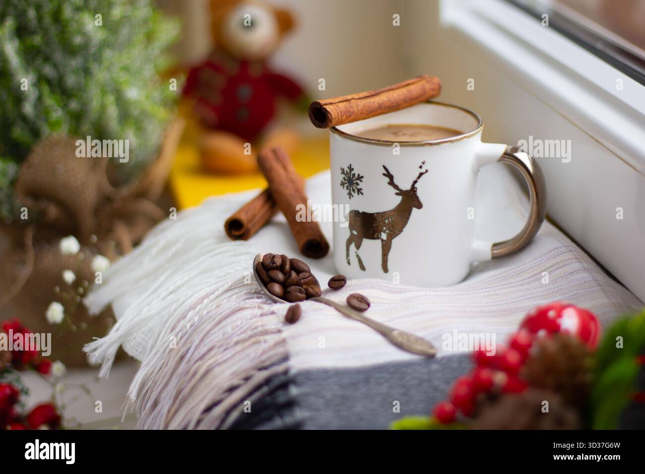Una scena accogliente presenta una tazza di caffè caldo adornata con bastoncini di cannella. I chicchi di caffè sono sparsi su una soffice coperta accanto alle decorazioni stagionali, c Foto Stock